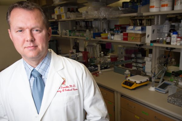 A photo of Dr. Nathan Sandbo standing in front of a research lab bench.